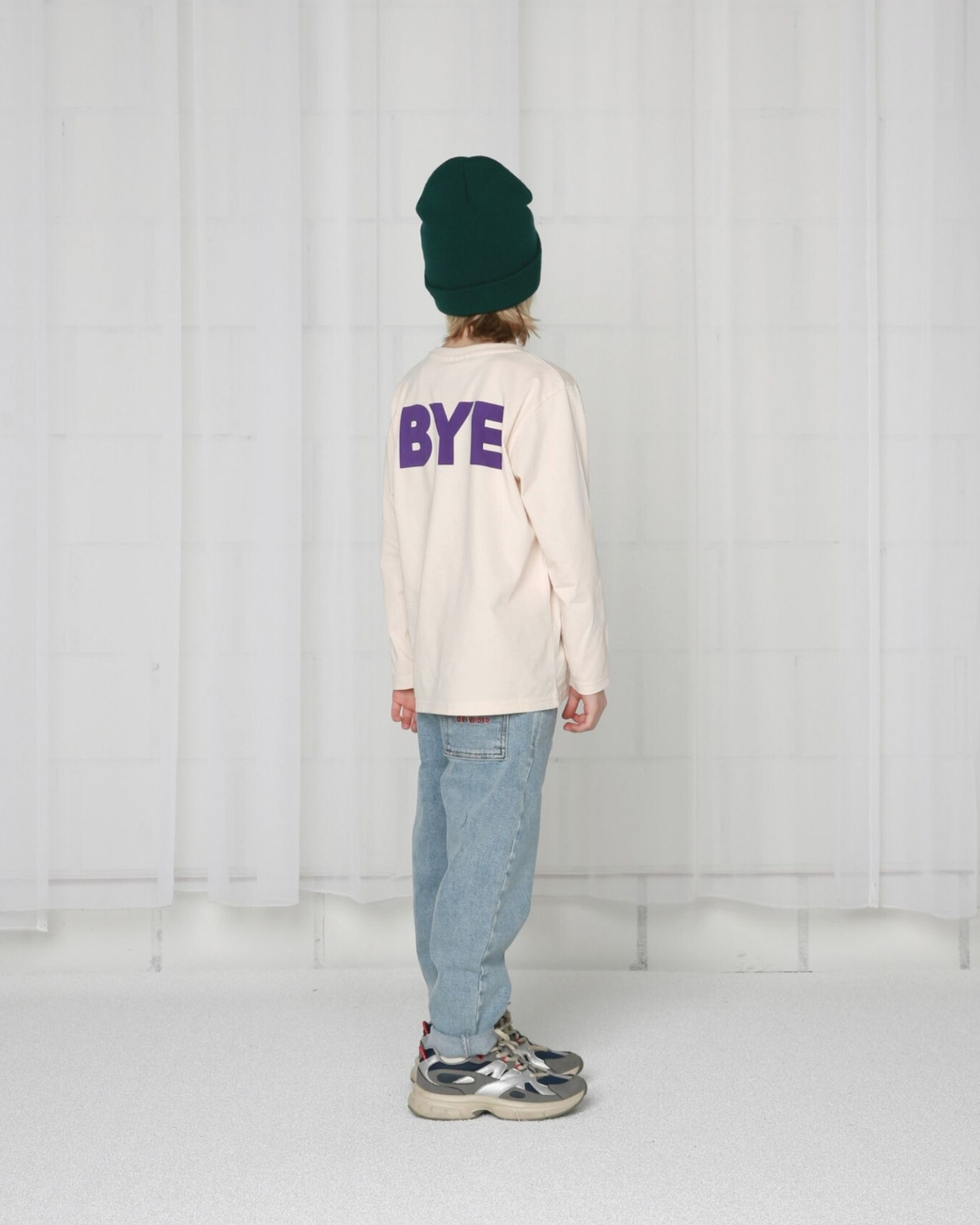 Boy model wearing the T-Shirt with long sleeves in colour Crystal Gray with caption BYE in dark purple from the rear.