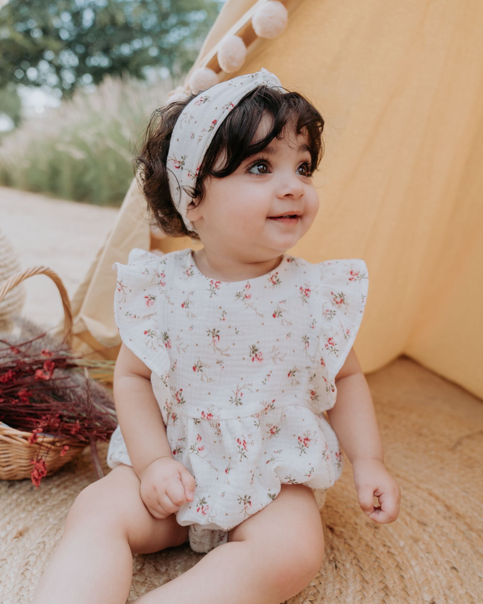 Baby Girl wearing the Rose Onesie in Wildflower Muslin Cotton. The romper is combined with the Twisted Headband in Wildflower Muslin Cotton.