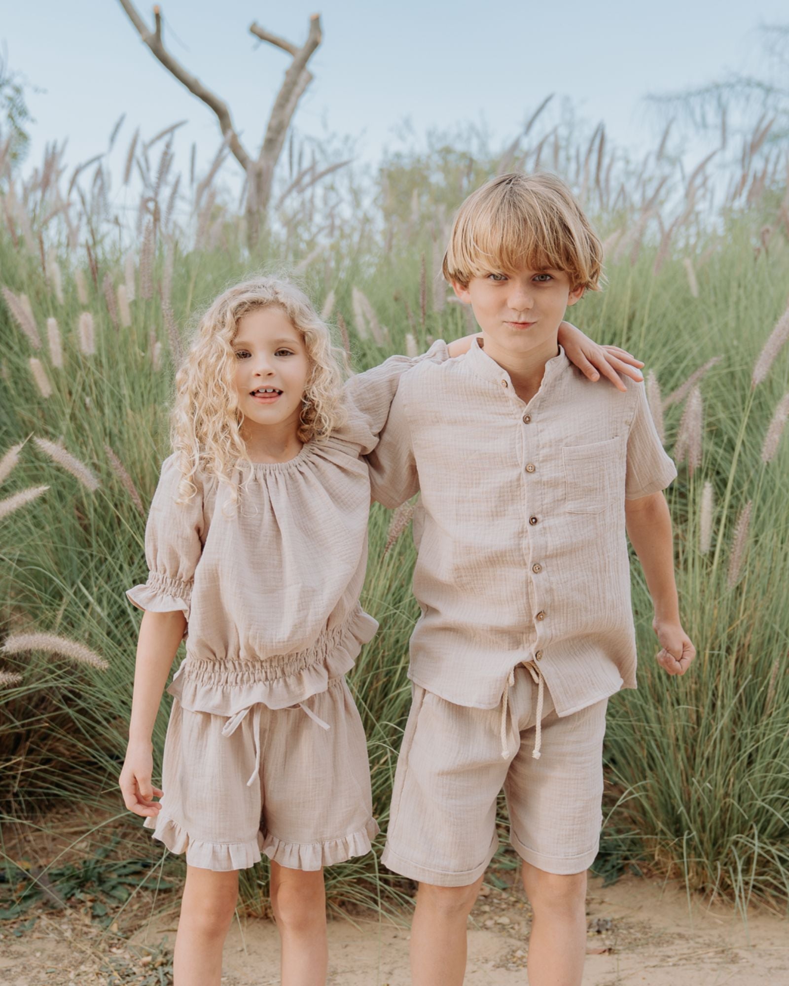 Girl model wearing the Alexa Top in Beige Muslin Cotton, combined with the Charlene Shorts in Beige Muslin Cotton. Twinning boy model wearing the Raphael Shirt in Beige, combined with the Julian shorts in Beige Muslin Cotton.