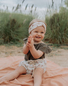 Baby Girl model wearing the Lilou Top in Soft Khaki Green Muslin Cotton with the Bloomers in Wildflower Muslin Cotton, combined with the Twisted Headband in Wildflower Muslin Cotton.